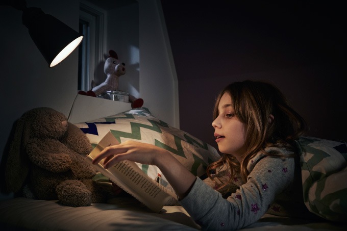 A girl wearing pyjamas lying in bed at night reading a book using a night light.