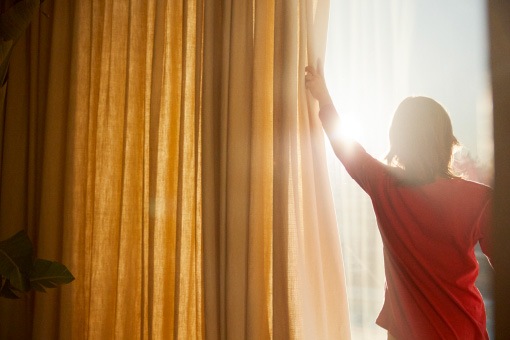 A boy wearing a red t-shirt opening up yellow curtains to let the sun in.