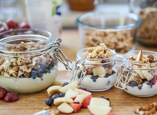 Three glass jars filled with yoghurt, granola, fresh fruit and nuts on a wooden countertop.