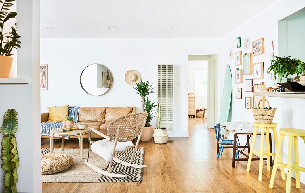 A living room with leather sofa, rattan rocking chair, hessian rug, wooden floors, and two yellow stools along a countertop.