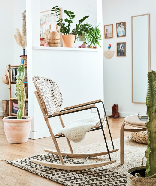 A living room with leather A rattan rocking chair on a hessian rug in a white room, with plants on a shelf and cacti in large pots on the wooden floor., rattan rocking chair, hessian rug, wooden floors, and two yellow stools along a countertop.