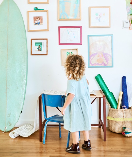 A small child stands in front of a small desk and chair, looking up at a wall of their paintings and family photographs next to a surfboard.