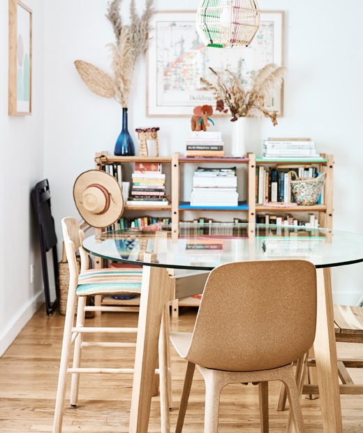 A dining area with glass-topped table and brown chairs, and a bookshelf against a white wall.