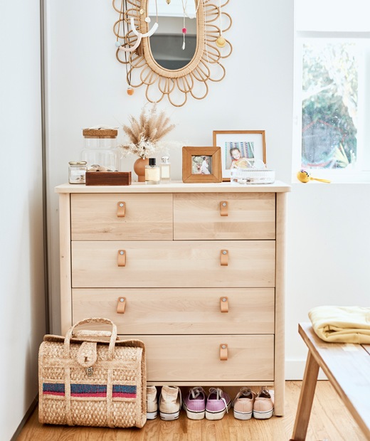 A light wood chest of drawers, with wicker-framed mirror on the white wall above, and a wicker bag and trainers underneath.