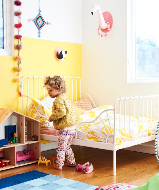 A small child in a room with yellow wall, white-framed bed with yellow and white bedding, and a wooden doll’s house.