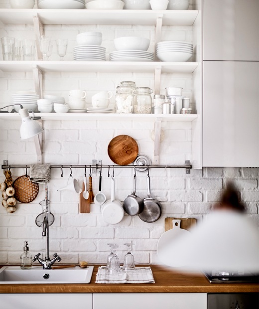 Barang pecah belah warna putih disimpan di rak terbuka di atas wastafel dapur dan meja dapur berbahan kayu.