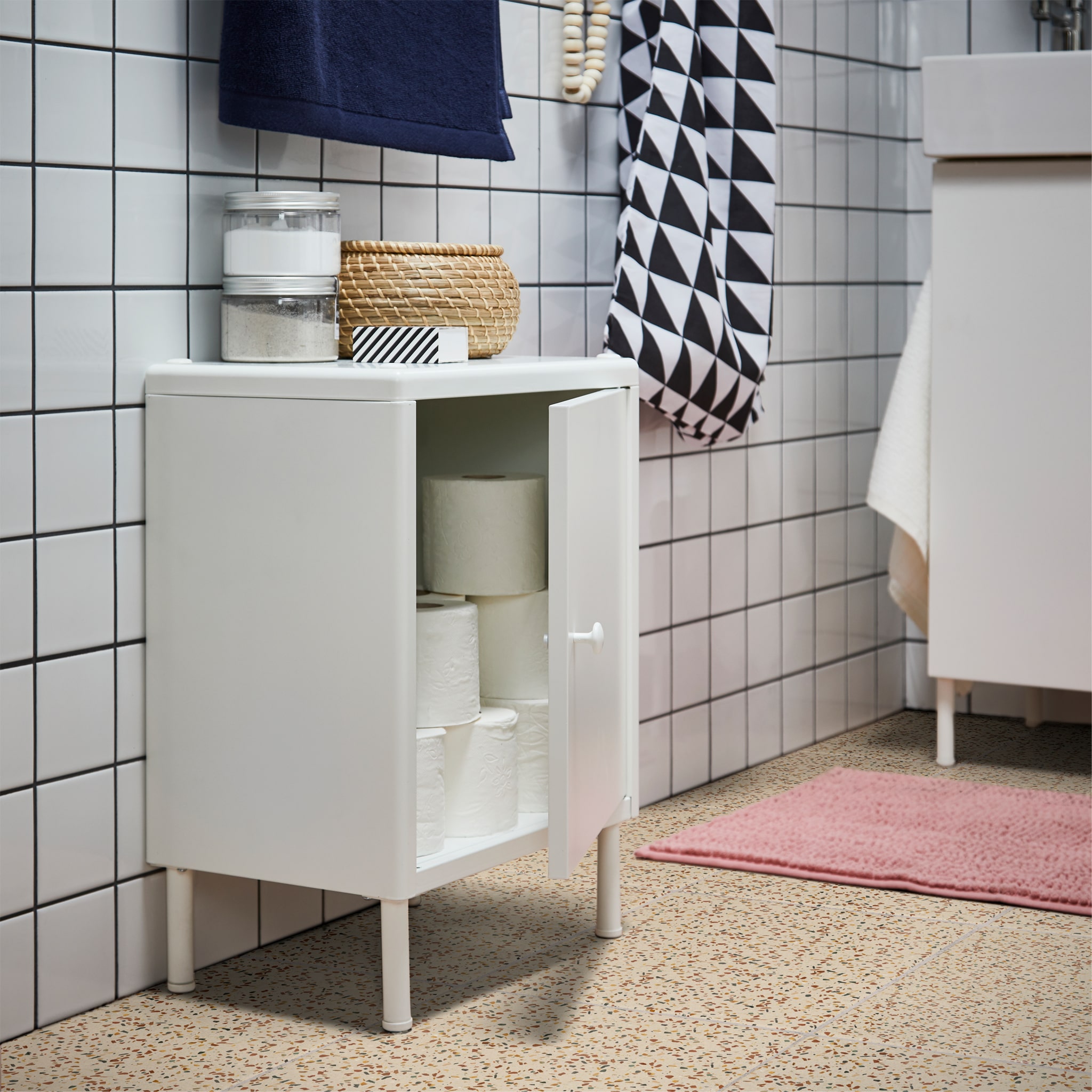 Small white cabinet with an open door, toilet paper inside. On top are transparent jars and a small seagrass box with lid.