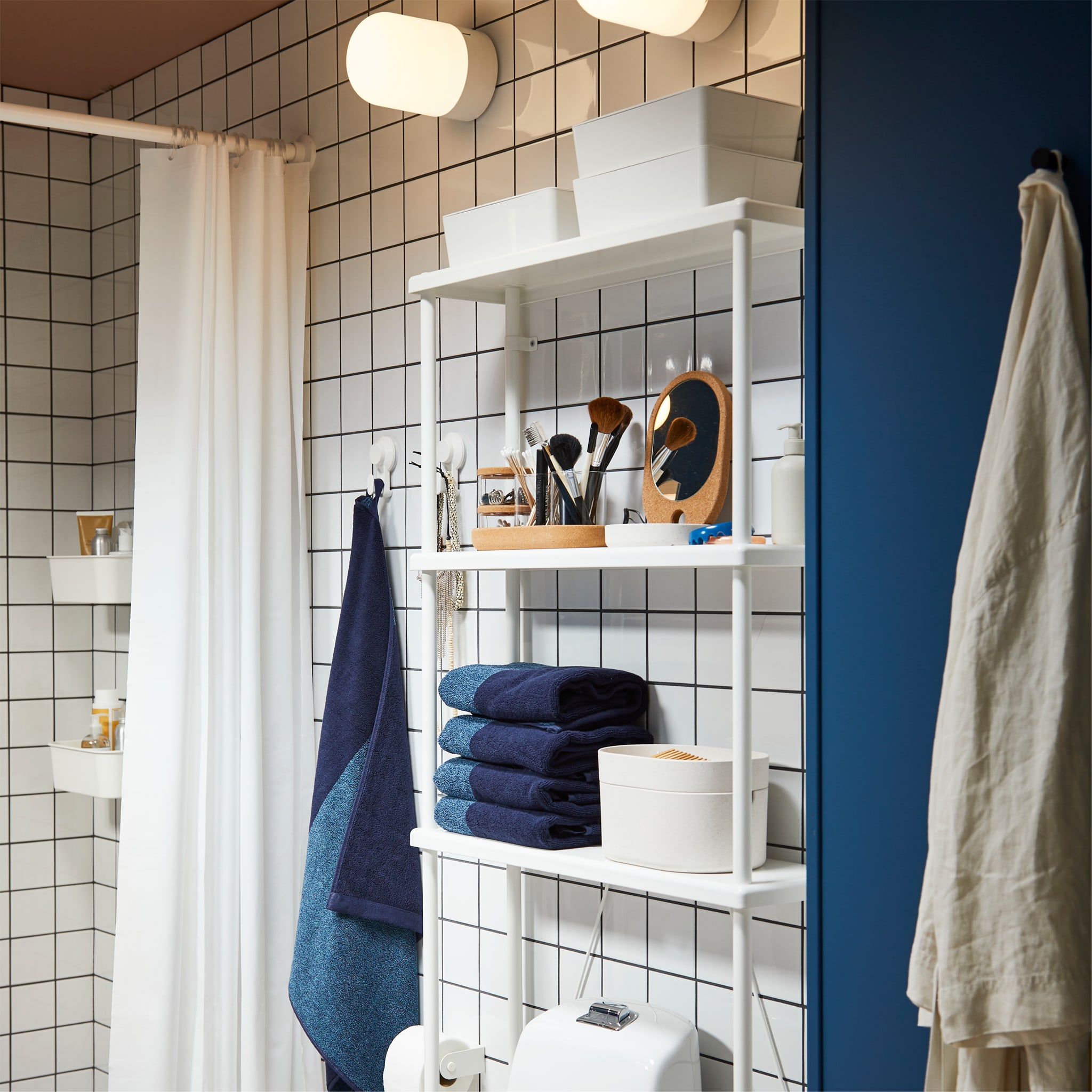 Bathroom with white shelves that store blue towels and white storage boxes above a toilet. White shower curtain hangs beside.