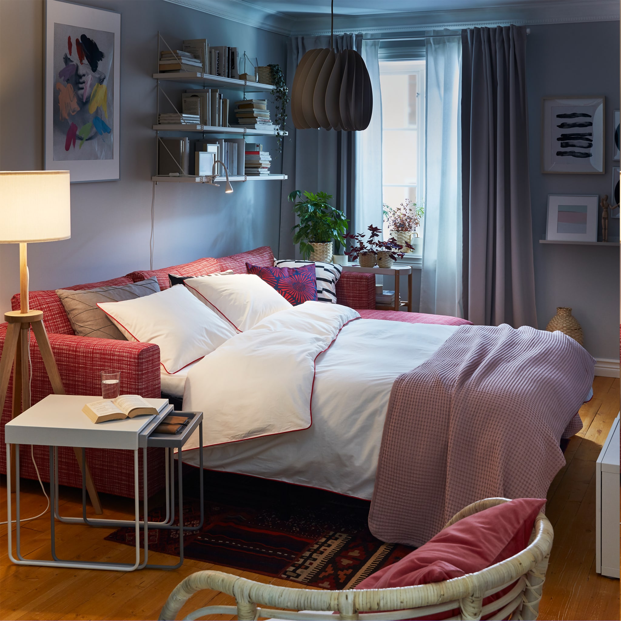  A red sofa-bed made as a bed with white bed textiles, a white nest of tables and a wooden and white floor lamp.