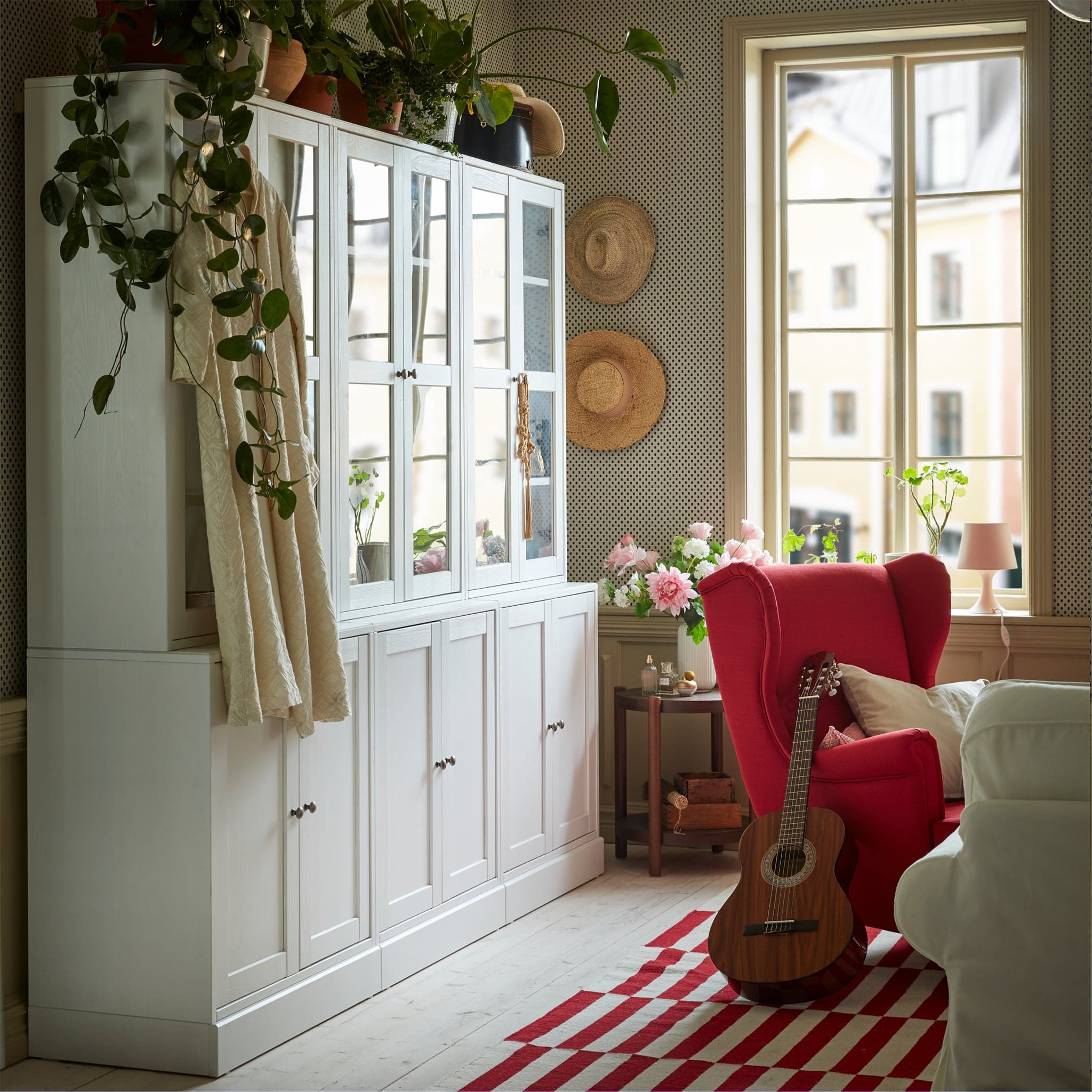  A white storage combination with glass doors in a red and white living room. Green plants are placed on top.