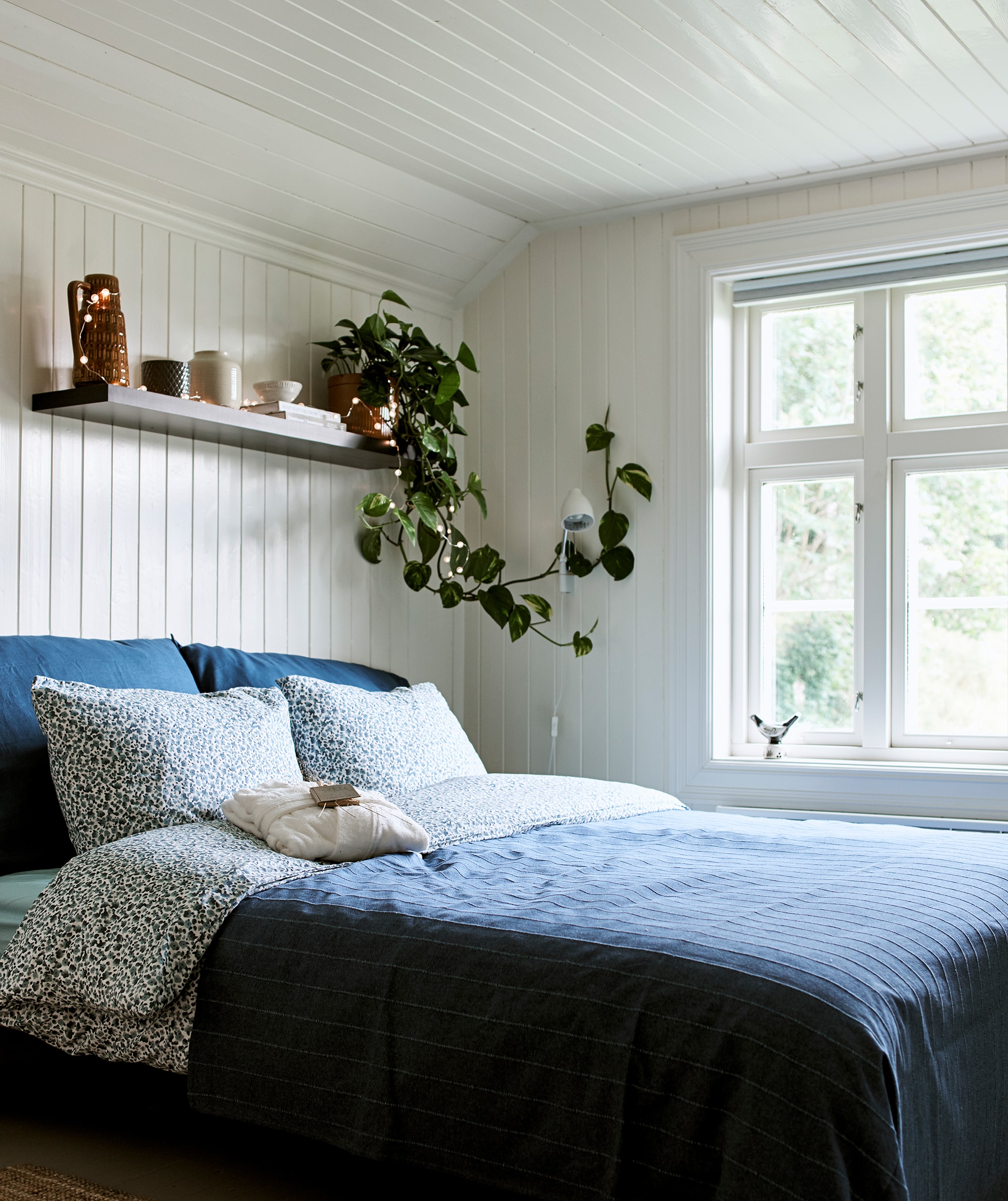 A farmhouse bedroom with white wood panelling and bed dressed in blue bedding with a wall shelf above to store and display.