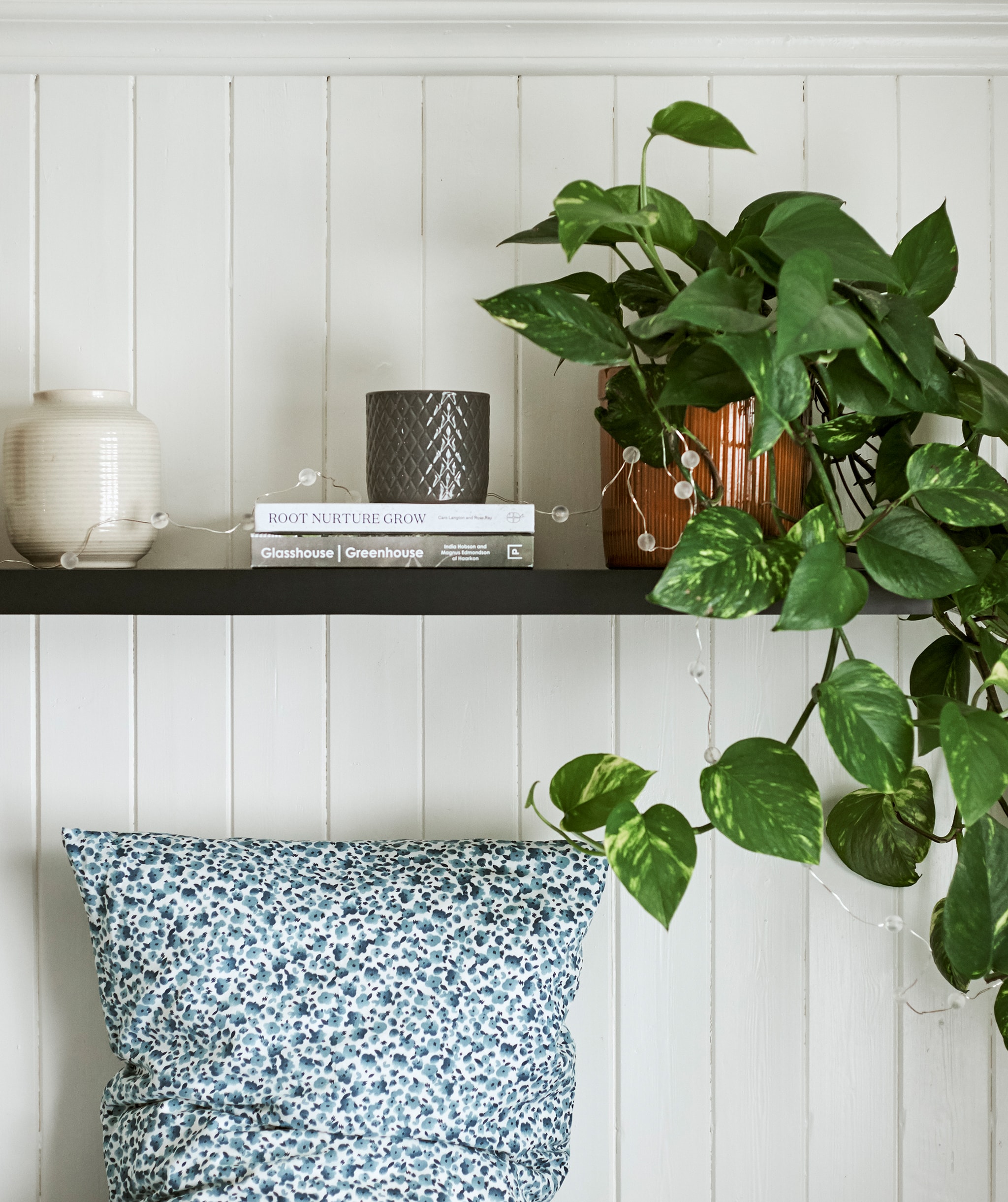 Sebuah tanaman ditampilkan menggunakan vas dan buku-buku di rak berwarna kayu gelap digantung di atas tempat tidur dengan bantal bermotif titik berwarna biru dan putih.