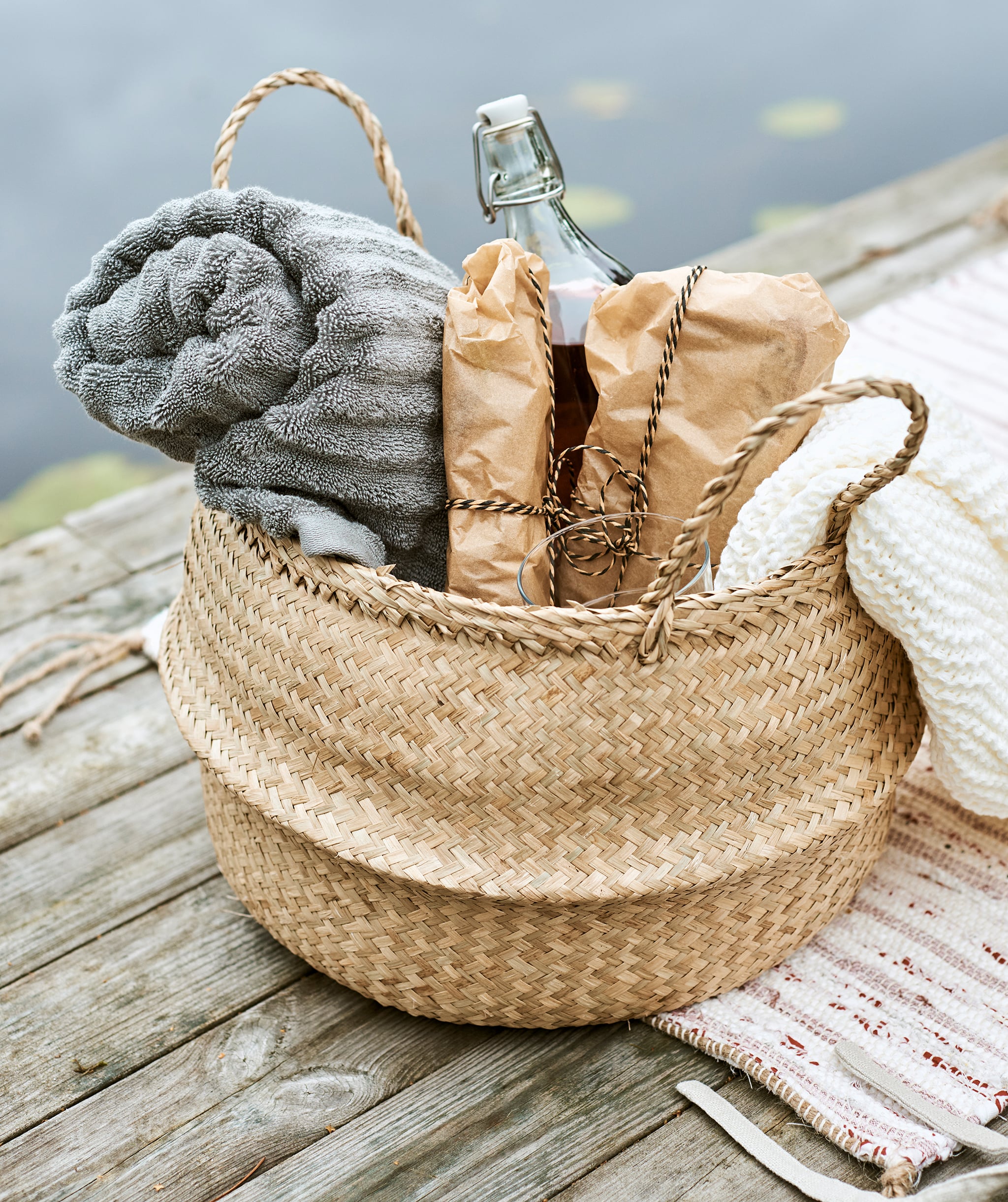 A woven basket packed for a picnic with rolled-up grey towel, sandwiches wrapped in paper, a cream throw and bottle of drink.