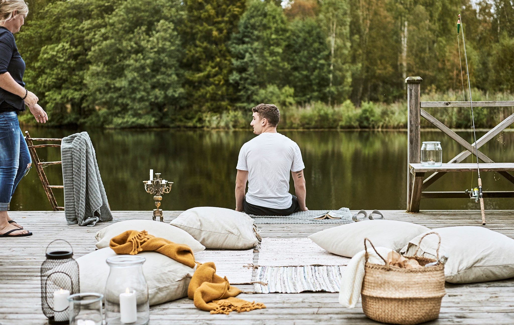A boy sits on the edge of a jetty next to a woman in flip-flops. On the deck are rugs, cushions, throws and a picnic basket.