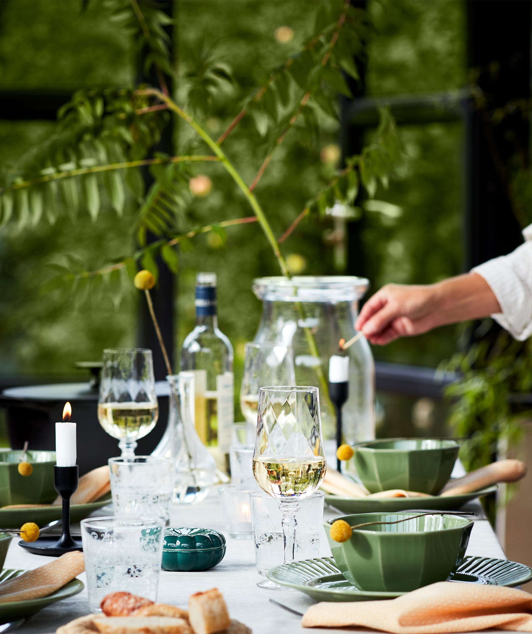 A well-laid table set for a dinner outdoors with green plates, wine glasses, candles and lots more. Shown with nature.