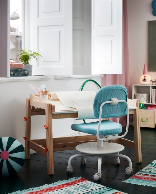 FLISAT children’s desk with a drawing paper roll stands by a window, and a turquoise children’s desk chair stands beside.