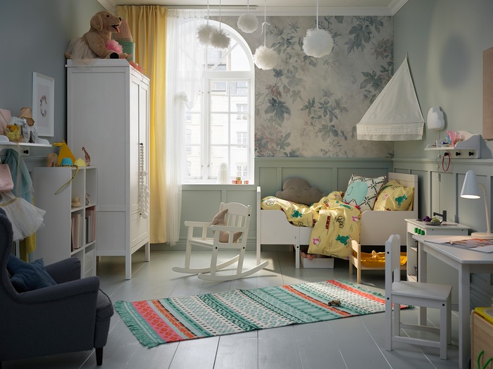 A children’s room with a bed, bed canopy, wardrobe, table and chair in white. A colourful rug lies across the floor.