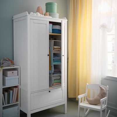 A rocking-chair and a wardrobe stand by a window with yellow curtains. One wardrobe door is open, folded clothes are inside.