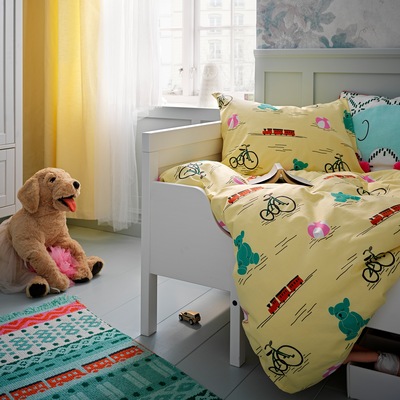 Yellow KÄPPHÄST bed linen with toy print in a white children's bed. A colourful rug and a soft toy dog on the floor beside.