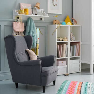 STRANDMON children’s armchair in dark grey stands next to a white shelf unit that stores books and toys.