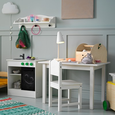 A children’s chair and a children’s table with a table lamp and crafts on top, and they stand next to a white play kitchen.