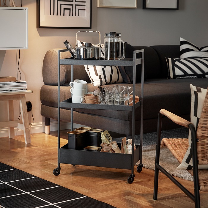 A black NISSAFORS trolley with a kettle, mugs and tea bags on its shelves is standing next to a sofa and armchair.