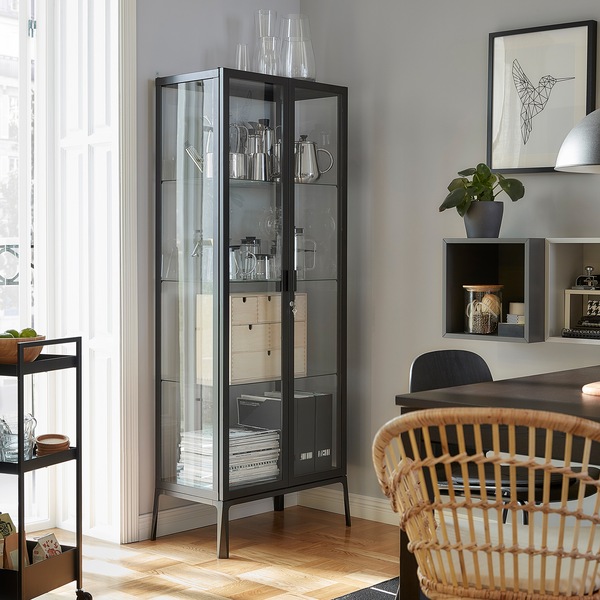 A MILSBO glass-door cabinet stands next to a balcony door. Decorative items, magazines and magazine files are stored inside.