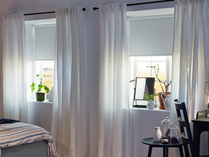 AINA curtains in white are hanging on black curtain rods in two bedroom windows where also white roller blinds are mounted.