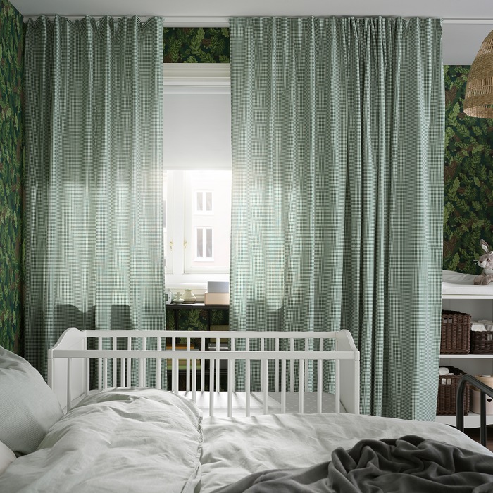 A bed and cot stand by a window with green/white curtains and a white block-out roller blind that covers half the window.