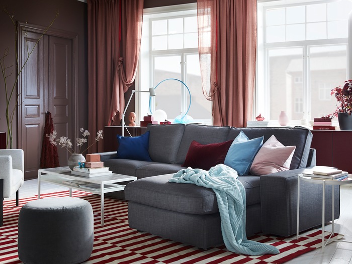 A living room with brown-pink curtains, a red/white rug, a grey sofa and wall-mounted cabinets with red high-gloss doors.