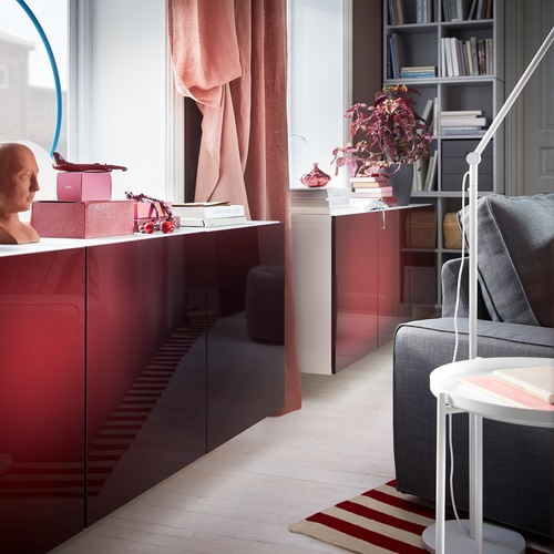 A white BESTÅ cabinet combination with high-gloss doors in dark red-brown is wall-mounted under a window.