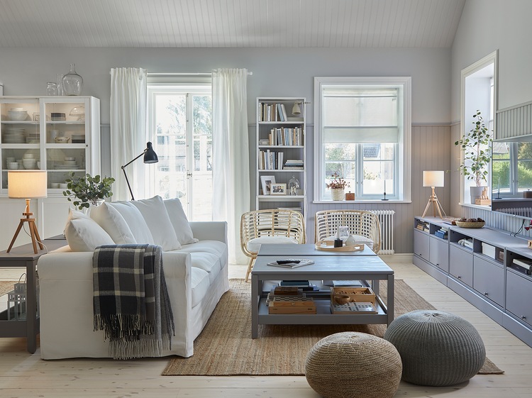 A traditional living room with a white sofa, a jute rug, two grey coffee tables, a grey TV bench and two armchairs in rattan.