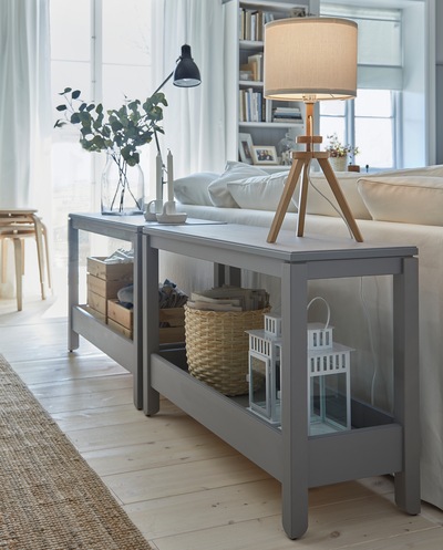 Two grey console tables are placed behind a sofa and serve as decorative room dividers. Two table lamps are placed on top.