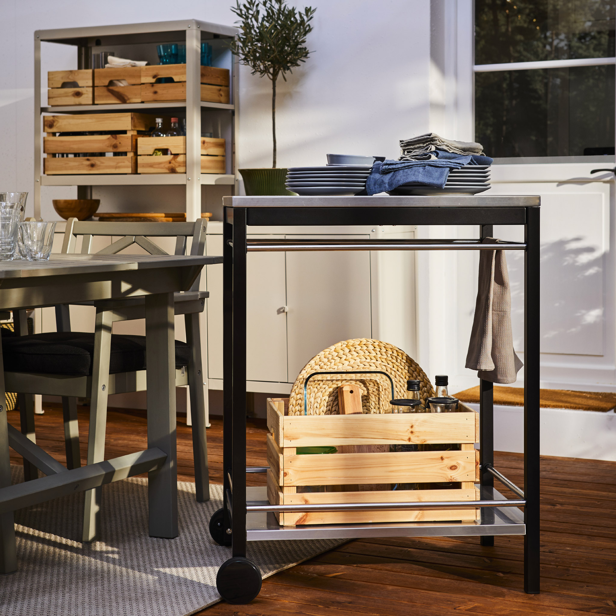 A trolley on castors in black/stainless steel holds dinnerware, towels and more, and it’s standing by a dining table.