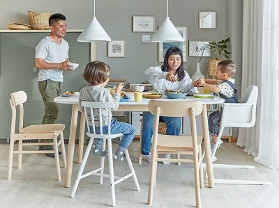 A family in a dining room. The mother and two children sit by the dining table, and the father brings a bowl to the table.