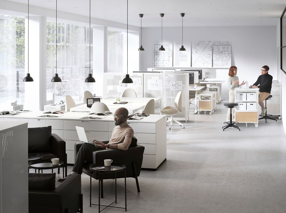 An open plan office with two breakout zones. A man works in a KOARP armchair and two people chat over a BEKANT shelving unit.