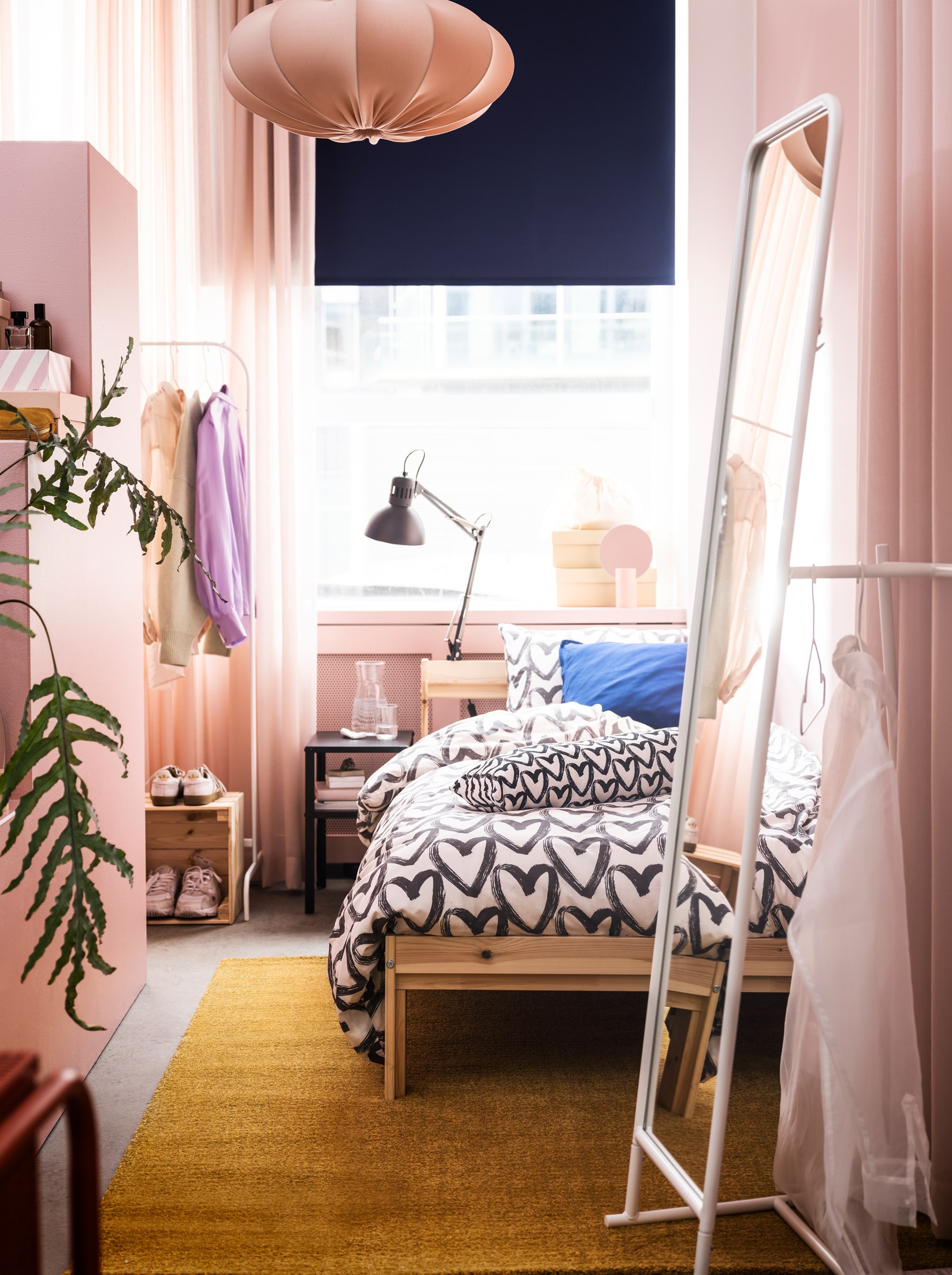 A narrow room in a rosy colour scheme with a NEIDEN bed, a tall KNAPPER mirror and ample room to store and display clothes.