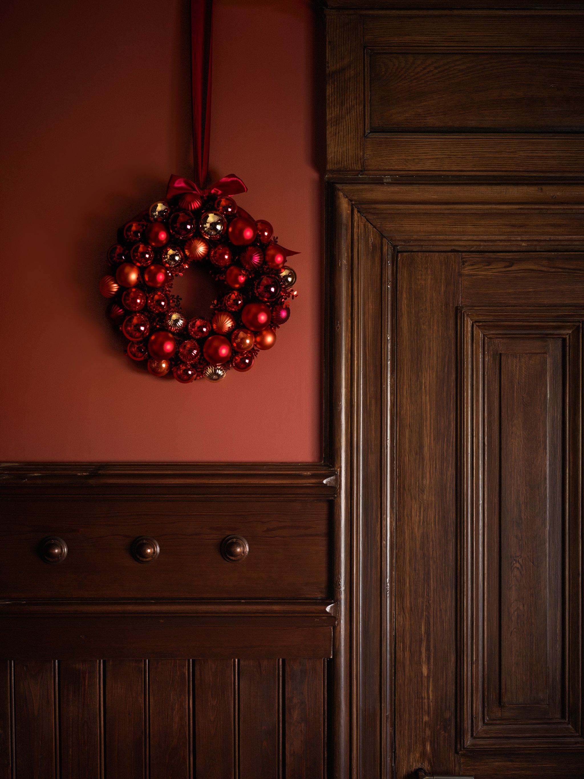 A red wreath of shiny baubles hangs from a red painted wall next to a large dark wooden door with ornate mouldings.