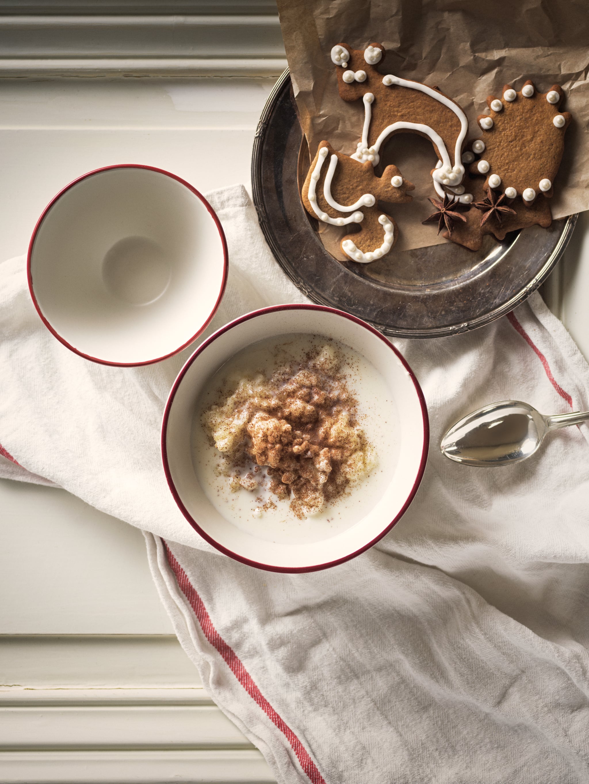 Two white VINTER 2020 bowls with red rims, one full of milky porridge, stand on a table next to a plate of gingerbread.